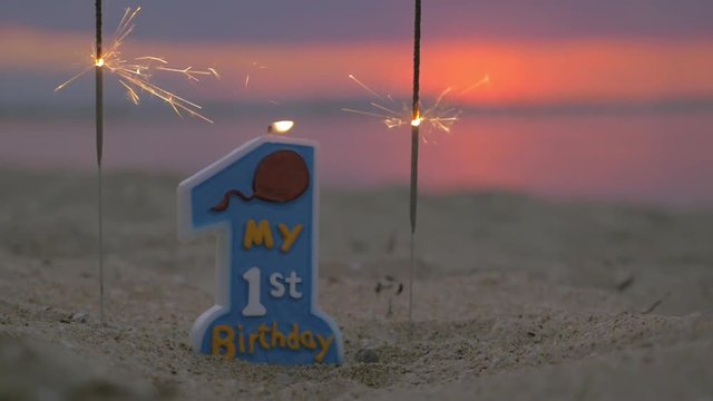 Slow motion close-up shot of burning one year old candle and sparklers on the beach at sunset. Baby boy birthday