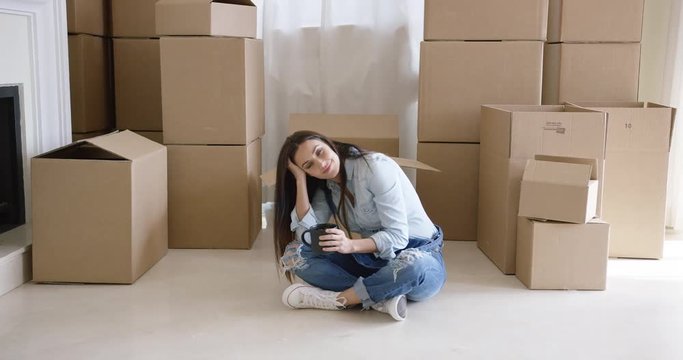 Tired young woman taking a break from moving house sitting cross-legged on the floor with coffee surrounded by boxes