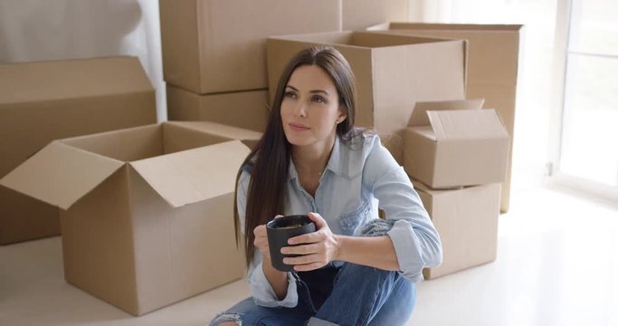 Thoughtful young woman squatting on the floor with a cup of coffee contemplating her new house surrounded by brown boxes