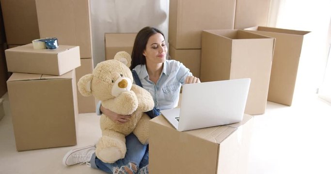 Young woman moving house with her large cuddly plush teddy bear sitting o the floor typing on a laptop balanced on a brown packing carton