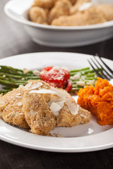 Parmesan Crusted Chicken Tenders with Roasted Sweet Potato and Roasted Asparagus topped with Campari tomatoes, Feta Cheese, Olive oil, and salt on a dark wooden background side view