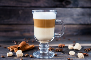 Hot latte macchiato coffee with tasty foam in tall clear glass on dark wooden table serving with cinnamon, cane sugar and roasted coffee beans.
