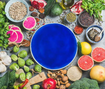Clean eating concept over grey background with dark blue plate in center, top view, copy space. Vegetables, fruit, seeds, cereal, beans, spices, superfoods, herbs for vegan, raw diet, gluten free diet