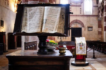 Ancient latin manuscript with religious chants and music sheets over a bookstand in the central nave of a romanesque church in an italian medieval abbey - obrazy, fototapety, plakaty