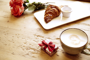 Fototapeta na wymiar Present box, rose flowers, fresh croissant, coffee on wooden table. Romantic breakfast for Valentine's Day celebrate.