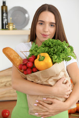 Young caucasian woman in a green apron is holding paper bag full of vegetables and fruits while smiling in kitchen