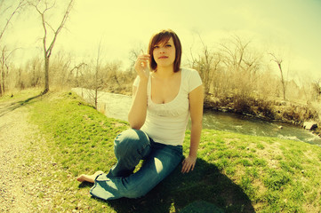 Young woman sits on the grass barefoot in a low-cut white t-shirt and jeans smoking a cigarette next to the river and trees in the spring. Girl smokies cigarette, looking at the camera. Filter effect.