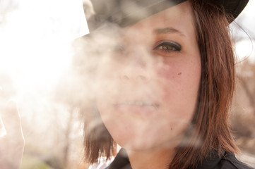 Cloud of backlit cigarette smoke obscure part of the model's face.  Cute white girl smokes a cigarette outside.  Black hat and black best.  Smoking outside.