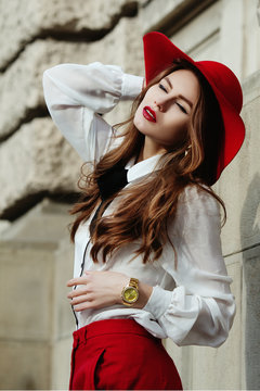 Outdoor waist up portrait. Young beautiful fashionable woman posing on street. Model closed her eyes. Lady wearing stylish red, white clothes, hat, wrist watch. Female fashion concept. City lifestyle