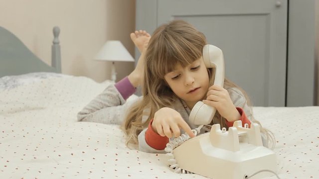 A pretty little girl lying on her bed, having a conversation on a vintage rotary phone.
