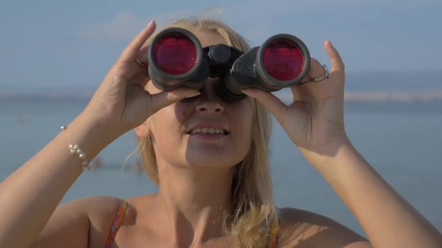 View of blond woman watching in binoculars with red glasses on landscape against blurred blue sea background