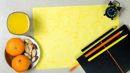 Black notebook, sheet of handmade yellow paper, color pencils, clock, glass of orange juice and healthy snack on light wooden background. Colorful work space. Desk dining.Top view. Ratio 16:9.