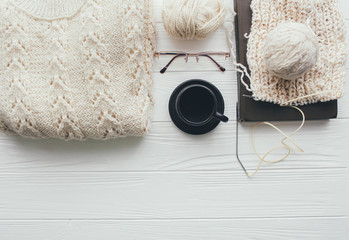 Knitted white sweater , needles , ball of yarn  coffee cup,glasses and book on   white wooden background