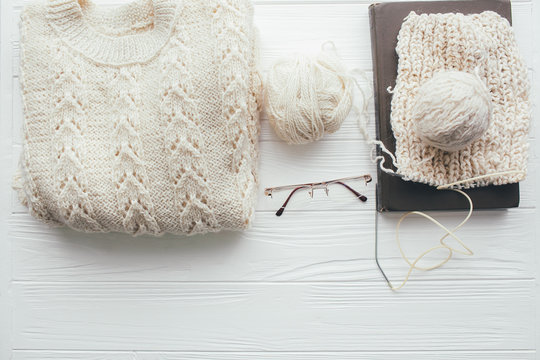 Knitted white sweater , needles , ball of yarn  coffee cup,glasses and book on   white wooden background