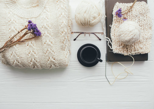 Knitted white sweater , needles , ball of yarn  coffee cup,glasses and book on   white wooden background
