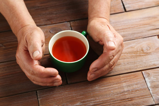 Hands of old senior with cup of tea on a wooden table