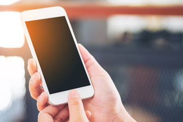 Mockup image of hands holding and touching white mobile phone with blank black screen in modern cafe