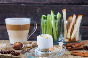 Soft Boiled egg with small spoon, coffee latte, sugar slices, cucumbers, bread for breakfast on old dark wooden table. Healthy food. Retro newspaper and cinnamon for decoration. Low key dark photo.