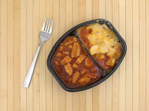 Microwaved TV dinner of chicken chunks in barbecue sauce plus potatoes atop a wood place mat with a fork to the side.