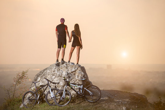 Rear view sports couple standing on a rock holding hands enjoying the evening scenery. Their sports bikes next to stone. Blurred background with copy space