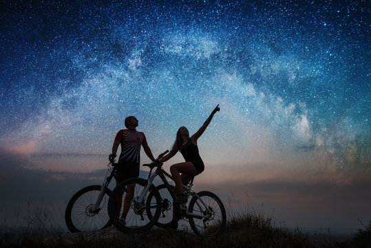 Sporting couple with mountain bikes on the hill under night starry sky. Woman shows man at the stars. Bottom view. Night landscape with colorful Milky Way.
