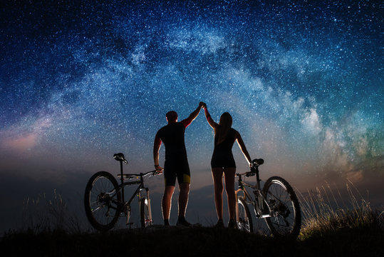 Night starry sky over cyclists on the hill. Guy and the girl with mountain bikes keep the hands lifted upwards to sky. Night landscape with colorful Milky Way. Bottom view