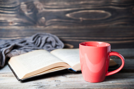 selective focus photo of grey cozy knitted scarf with cup of coffee or tea and open book on a wooden table.