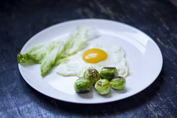 Obraz na płótnie Canvas fried egg with cucumbers, olives, and brussels sprouts on a plate with fork and knife on a dark wooden background