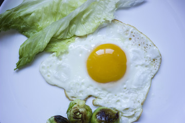 fried egg with cucumbers, olives, and brussels sprouts on a plate with fork and knife on a dark wooden background