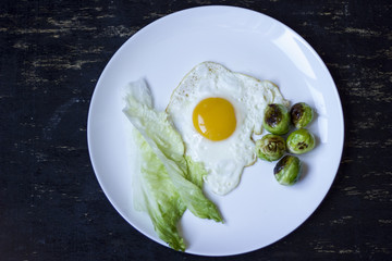 fried egg with cucumbers, olives, and brussels sprouts on a plate with fork and knife on a dark wooden background