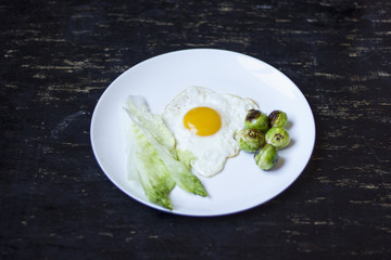 fried egg with cucumbers, olives, and brussels sprouts on a plate with fork and knife on a dark wooden background