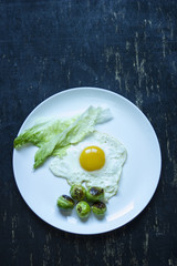 fried egg with cucumbers, olives, and brussels sprouts on a plate with fork and knife on a dark wooden background