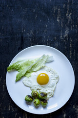 fried egg with cucumbers, olives, and brussels sprouts on a plate with fork and knife on a dark wooden background