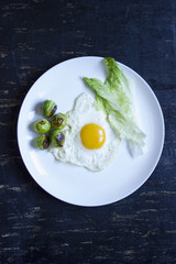 fried egg with cucumbers, olives, and brussels sprouts on a plate with fork and knife on a dark wooden background