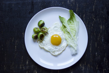 fried egg with cucumbers, olives, and brussels sprouts on a plate with fork and knife on a dark wooden background