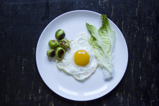 Fried Egg With Cucumbers, Olives, And Brussels Sprouts On A Plate With Fork And Knife On A Dark Wooden Background