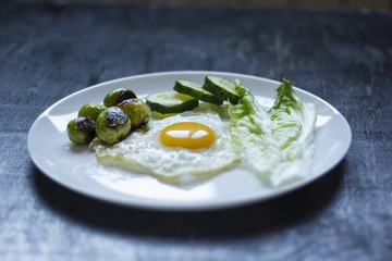 fried egg with cucumbers, olives, and brussels sprouts on a plate with fork and knife on a dark wooden background