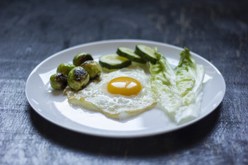 fried egg with cucumbers, olives, and brussels sprouts on a plate with fork and knife on a dark wooden background