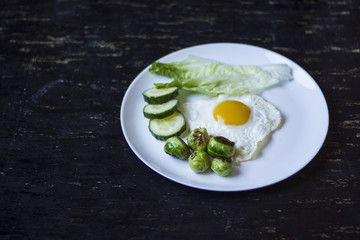 fried egg with cucumbers, olives, and brussels sprouts on a plate with fork and knife on a dark wooden background