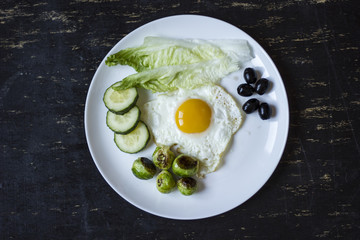 fried egg with cucumbers, olives, and brussels sprouts on a plate with fork and knife on a dark wooden background
