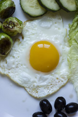 fried egg with cucumbers, olives, and brussels sprouts on a plate with fork and knife on a dark wooden background