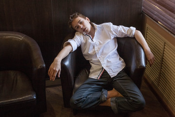 Boy in white shirt sitting in a chair. young boy imposingly placed in a leather couch. boy in shirt and jeans. Teenager relaxing in a cafe
