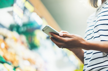 Young woman shopping healthy food in supermarket blur background. Female hands buy products tomato using smartphone in store. Hipster at grocery using smartphone. Person comparing price of produce