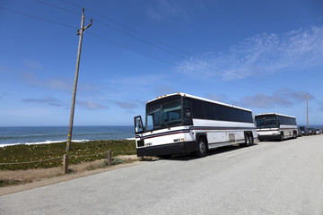 Obraz na płótnie Canvas Two white parked tour buses along California Pacific coast with ocean and blue sky.