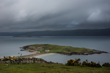 North Coast, Scotland - June 6, 2012: Small green island in Loch Eriboll, connected with a short dam, and featuring a white fisherman house. Cloudy foggy sky, bluish water, mountainous background.