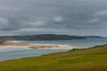 North Coast, Scotland - June 6, 2012: large sandbank blocks partly the mouth of Naver River under gray cloudy sky. Azure blue water, green foreground, mountainous background.