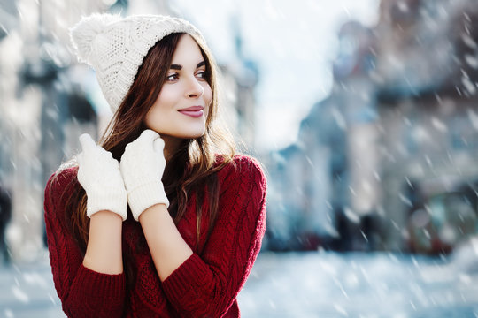 Outdoor waist up portrait. Young beautiful happy smiling girl walking on street. Model looking aside, wearing stylish sweater, hat, gloves. City lifestyle. Magic snowfall. Empty, copy space for text