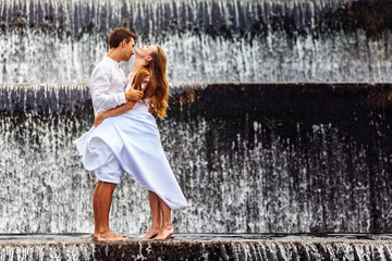 Happy family on honeymoon holidays - married loving couple hugging, kissing with fun under falling water in cascade waterfall pool. Active lifestyle, people travel on summer vacation on Bali island.