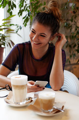 Handsome happy Latin young woman with beautiful smile in the cafe near the window with coffee smiling, cute hipster female person using mobile phone sitting in cozy coffee house with friends