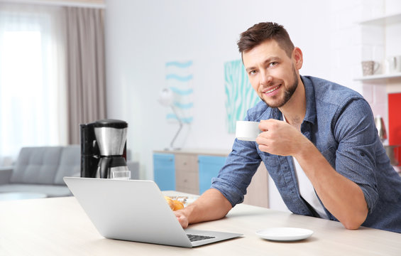 Handsome young man drinking coffee while working with laptop in kitchen